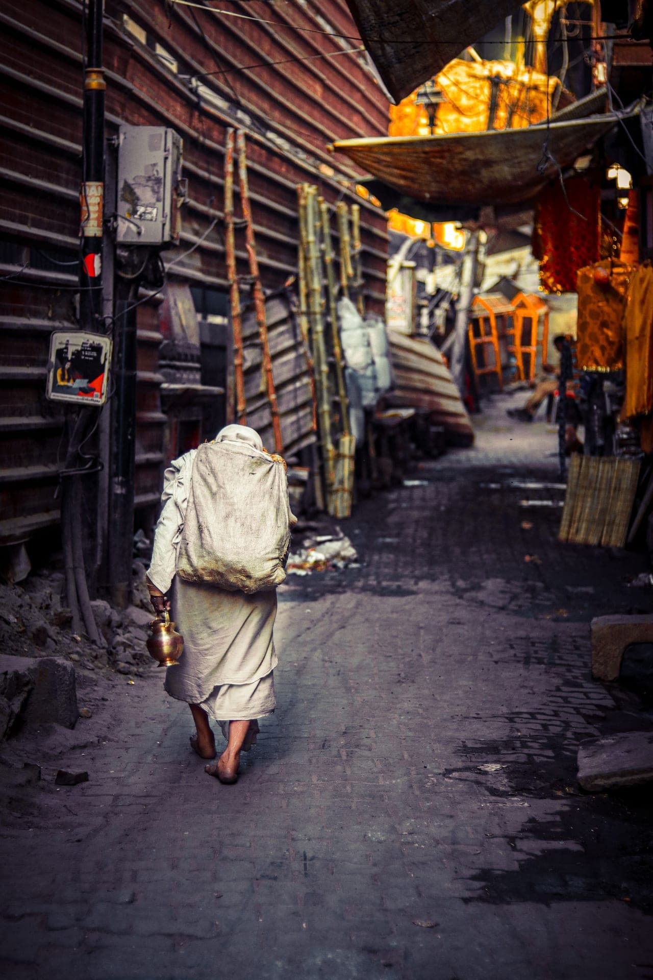 A lane in old Varanasi, a lone figure walking toward the market lights with a brass water-pot.
